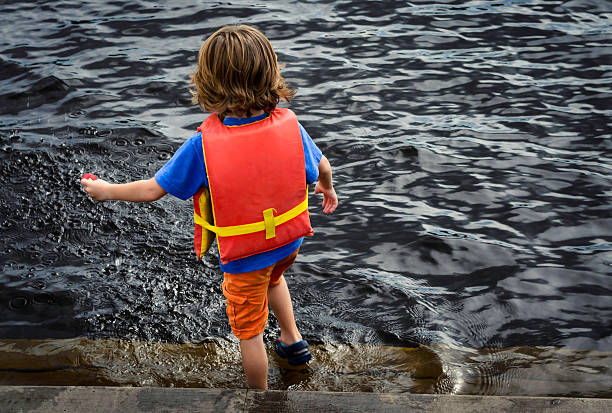 Young boy playing near the edge of a lake stepping into the water and wearing a red life jacket. 