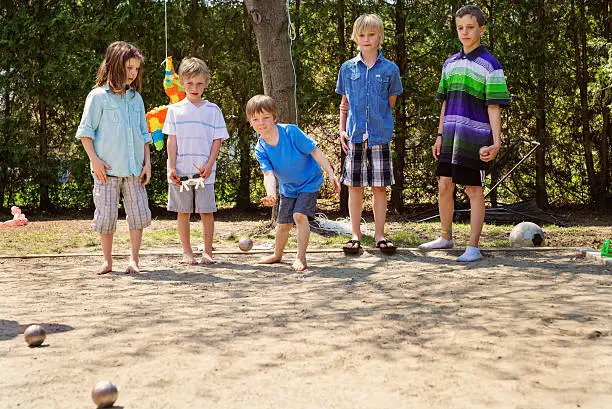 Little girl playing a ball game outdoors with the boys in a suburb backyard. She is dressed in unisex clothes, confortable for playing. They are playing petanque, a popular french game. This was taken on a hot and sunny spring day. Horizontal full length shot with copy space.