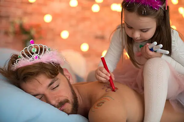 Cute daughter drawing butterfly and rainbow with felt pen on her sleeping father's back. Father is lying on stomach and wearing tiara.