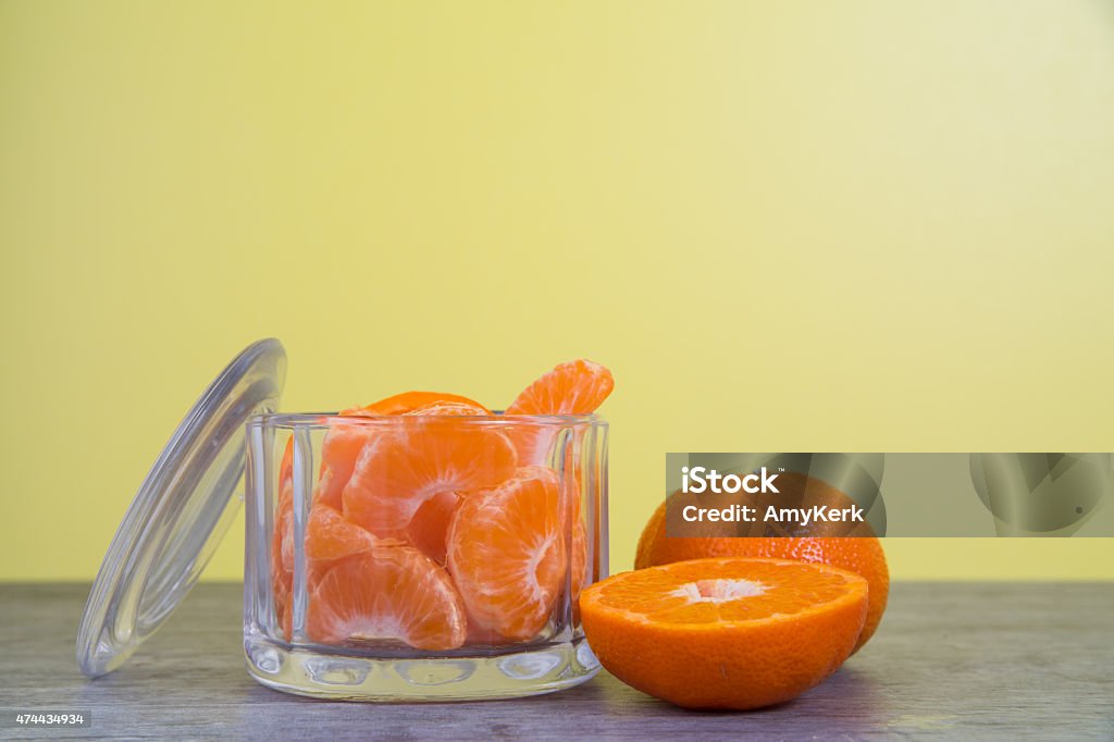 Mandarin oranges in a glass jar with a yellow background Mandarin orange slices in a glass jar sit on a rustic wooden table with a bright yellow background.  One whole orange sits behind a halved orange to the side of the glass jar. 2015 Stock Photo