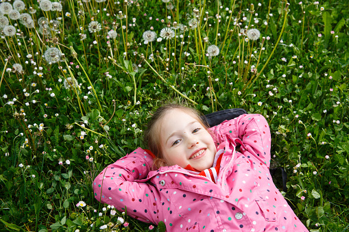 Beautiful little girl lying in grass, surrounded by dandelion seed heads, enjoying nature and its serenity. Natural childhood concept.