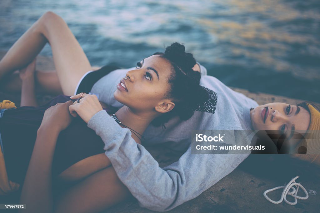 Teen grunge girl friends lying together alongside water Cropped image of mixed race teen girl friends in grunge styling lying alongside water together City Stock Photo