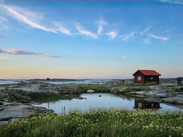 Small red wooden hut on a rocky skerry in the outer archipelago of Stockholm, Sweden. Grass in front and reflections in a small pond of water in front of the house.