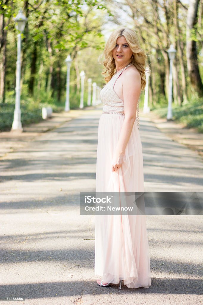 Blond woman in evening gown posing Beautiful blond curly woman wearing evening peach color gown posing outdoors. Fashionable and glamorous dress and style. Blond Hair Stock Photo