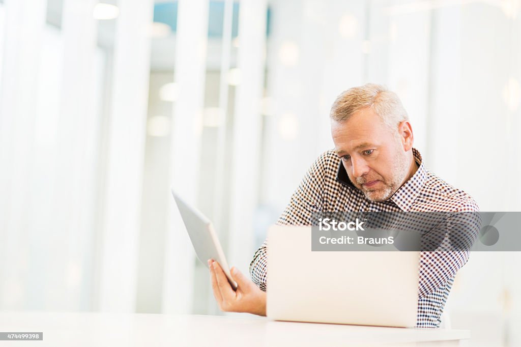 Multi-tasking businessman using wireless technology in the office. Busy mature businessman working in the office. He is talking on the phone and using laptop and touchpad. 2015 Stock Photo