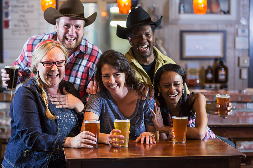 A multi-ethnic group of five friends drinking beer in a restaurant bar.  The women are sitting at a table holding their drinks, and two men wearing cowboy hats are standing behind them.  They are all smiling, laughing and looking at the camera.