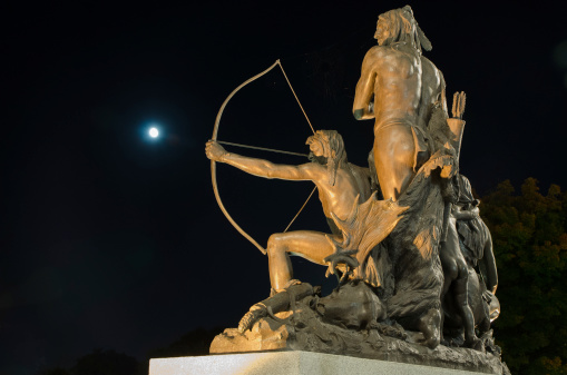 This a sculpture of an Indian family with one ready to shoot an arrow with his bow. Picture taken at night with full moon just in front of the arrow. 