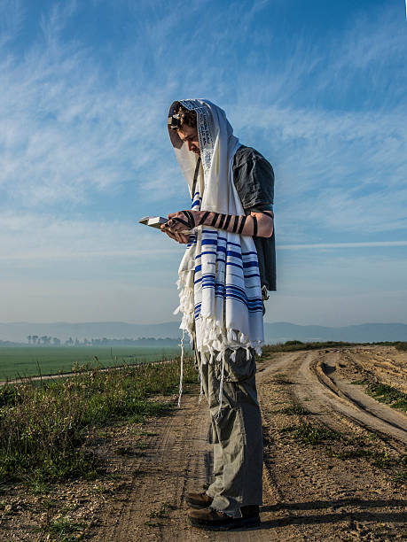 Color image depicting the rear view of a Jewish man wearing a blue skull cap (or Kippah) embroidered with the Star of David while sitting alone on a bench in the park. Focus on the man with the lush foliage of the park defocused beyond.