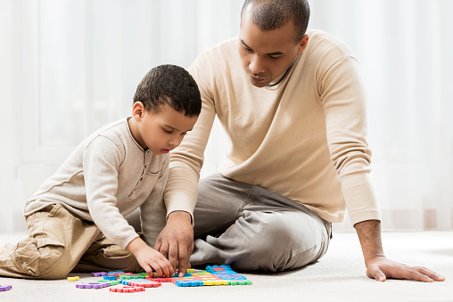 African American mid adult man and his little boy sitting on the carpet at home and playing with puzzles.