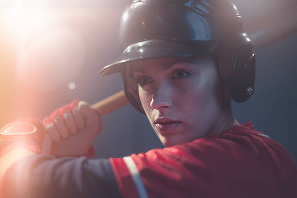 Female Baseball Superstar A very shallow depth of field close up portrait of a female baseball player, holding baseball bat behind her during a baseball game, ready to strike. The woman is wearing a safety helmet and generic red baseball uniform and gloves.  The image intentionally contains lots of lensflare.  baseball hitter stock pictures, royalty-free photos & images