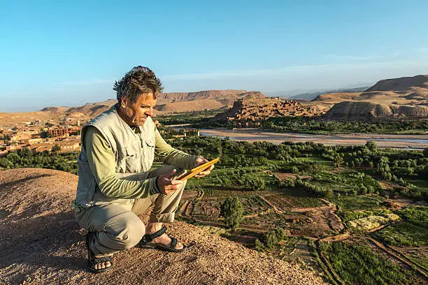 Mature man in natural colour clothes using his digital tablet while crouching on the hill. Behind him on the right the old fortified city (ksar) Aït Benhaddou, on the left the new city, central Morocco. This ancient city, giant fortification, is an example of earthen clay architecture. It is made up of six Kasbahs and nearly fifty ksars which are individual Kasbahs. Many films have been shot there.  It is a UNESCO World Heritage Site. Nikon D800, full frame, XXXL.