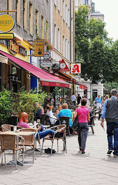 Berlin, Germany - August 3, 2011: People walking through Berlin Kreuzberg (Germany) at Mehringdamm, in Front small shops, restaurants. a man in chair reading magazine. Its a touristical magnet of multi-culture.
