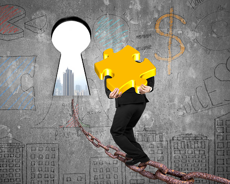 Man carrying golden jigsaw puzzle walking on old iron chain toward keyhole door, with urban scene view and business concept doodles wall background
