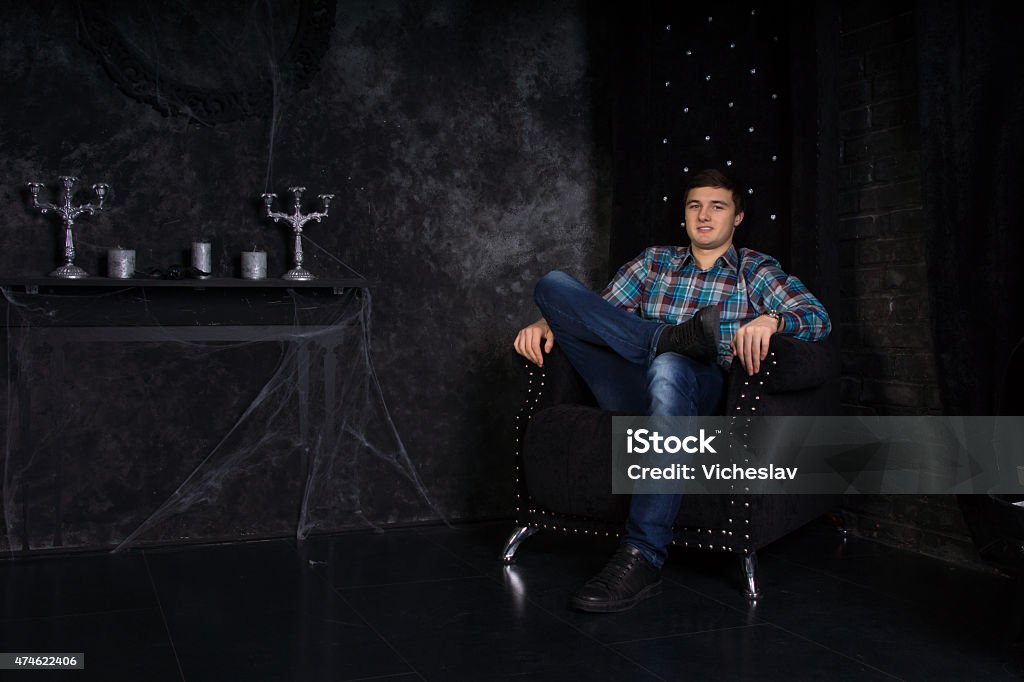 Man Sitting on High Back Chair in Haunted House Smiling Young Man Sitting with Legs Crossed in Plush Black High Back Chair in Eerie Haunted House Setting with Cobwebs Men Stock Photo