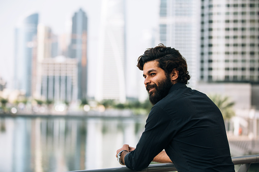 Young man with beard standing outside. He is wearing black shirt and looking at the view and smile