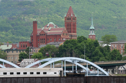 A small railroad engine, shunter, shifter or switcher, pulls a single passenger railroad car over the north branch of the Potomac river with the Allegany County Circuit Court building in the background in Cumberland Maryland.