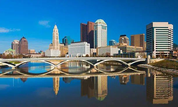 Downtown Columbus skyline and a modern bridge with the Scioto River with a reflection of the scene in the foreground.