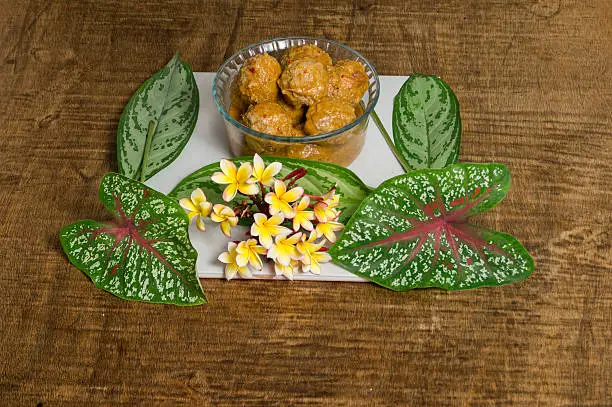 A crystal bowl of Mexican Meat Balls (albóndigas in Spanish). Cooked with chipotle chili. May flowers bouquet and green leaves with red and white stripes. Served over a wooden table.  The dish is called in México Albondigas al chiplote.