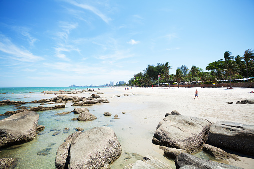 Hua Hin, Thailand - April 3, 2015: Capture of people on Hua Hin beach at sunny day. In foreground are rocks in sand and sea. View along beach to south. At right side are palm trees and resorts. In far background are hotels along coast.