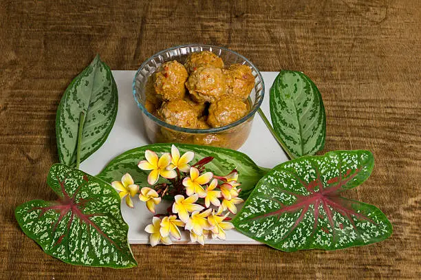 A crystal bowl of Mexican Meat Balls (albóndigas in Spanish). Cooked with chipotle chili. May flowers bouquet and green leaves with red and white stripes. Served over a wooden table.  The dish is called in México Albondigas al chiplote.