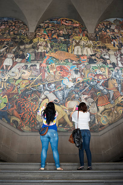 Mexico City, Mexico - October 28, 2014: Two women stand on steps at the National Palace in Mexico City, Mexico, using their smartphones to photograph a mural by Diego Rivera, one of several painted between 1929 and 1951. The open-air murals are one of the most visited attractions in Mexico City; entrance is free.