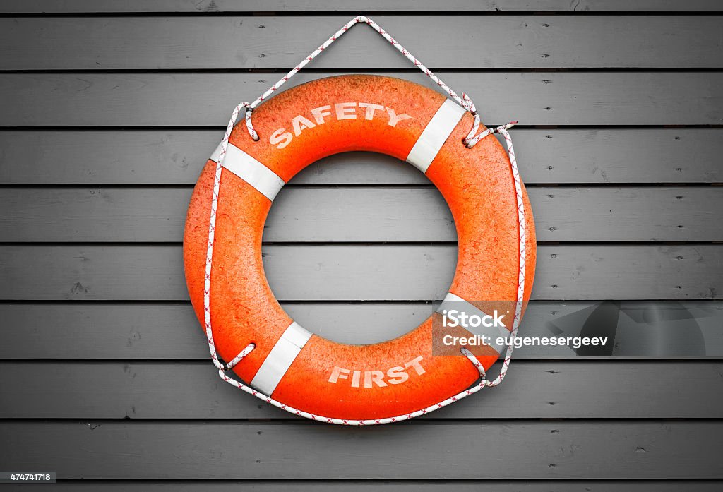 Safety first. Red lifebuoy hanging on gray wall Safety first. Red lifebuoy with text label hanging on gray wooden wall of a port building Buoy Stock Photo