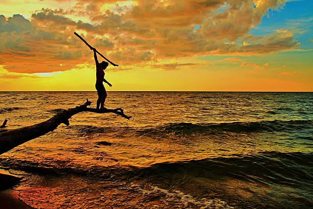 Hunt Silhouette of an adolescent balanced on a log preparing to spear a fish. spear stock pictures, royalty-free photos & images