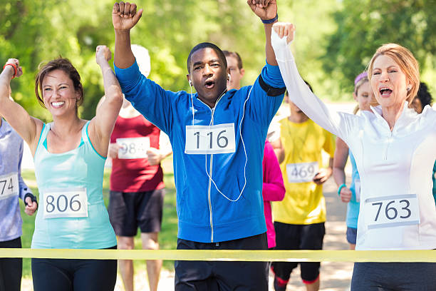 Diverse team of runners celebrating while crossing race finish line Mature Caucasian women and mid adult African American man are raising their hands in celebration as they cross yellow finish line tape. They are winning marathon or 5k race for charity. Runners are wearing athletic clothing and race contestant numbers. Other runners are behind them during race in park on sunny day. walking for a cause stock pictures, royalty-free photos & images