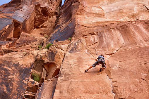Young man carefully scaling a red sandstone rock cliff with full climbing gear at the Wall Street area near Moab Utah, a haven for climbers.