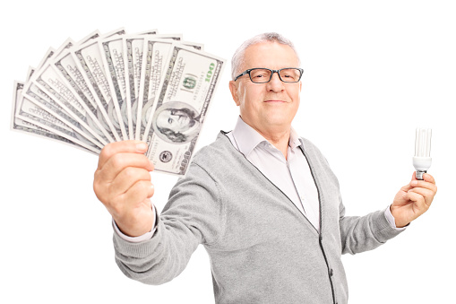 Economic senior holding an energy efficient light bulb and a stack of money isolated on white background