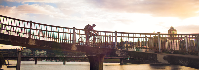 A panoramic view of a young man commuting in an urban city environment on his street bicycle pauses on a bridge to view whatever is below.  Dramatic sunset sky and clouds.  Vertical with copy space.