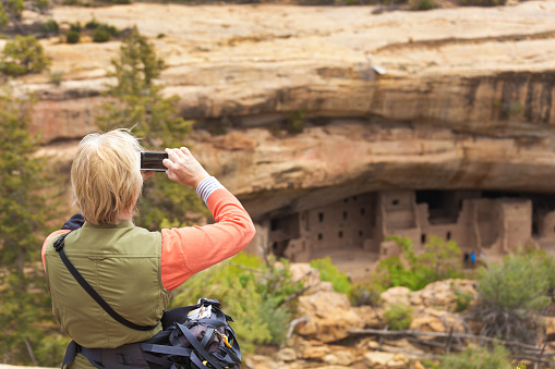 Woman tourist visiting and photographing the cliff dwelling site of the ancient pueblo tribe in the Mesa Verde National Park, Colorado, USA. The complex adobe cliff architecture was built by the southwest American ancient Pueblo, featuring housing, worship sites, storage and community space.