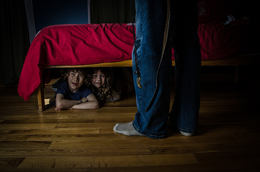 Two kids (brother and sister) are hiding under the bed with fear while the father is in front of the bed with his belt hanging besides him