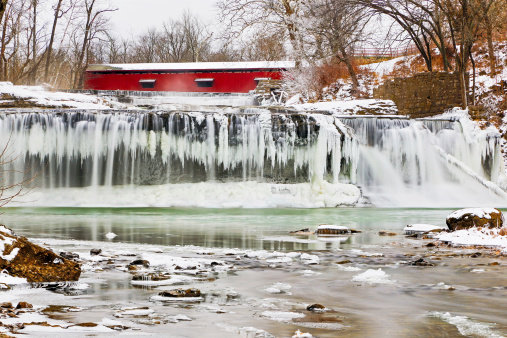 Whitewater flows over icicles at a frozen Upper Cataract Falls with a red covered bridge upstream. Photographed on Mill Creek near Cloverdale, Indiana.