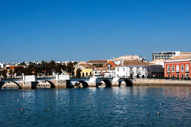 View across the Gilao river to the old bridge and town of Tavira in the Algarve province of Portugal. There is a large expanse of river in the foreground with some seagulls on the water and a large expanse of clear blue sky overhead for copy.