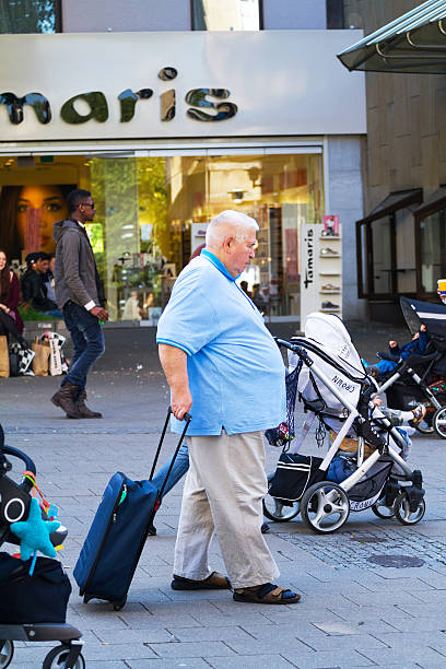 Essen, Germany - May 15, 2015: Sideview of a caucasian senior man walking with blue shirt and shopping trolley in pedestrian zone and shopping street of Essen. In scene are people and three baby buggies. In background a black man is walking.