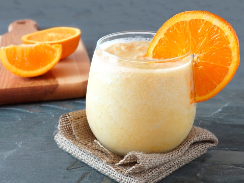 Healthy orange fruit smoothie in a stemless glass resting on burlap cloth with fresh orange slices in background