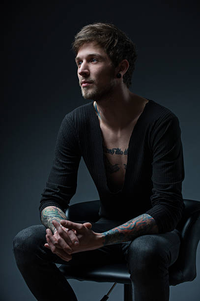 young guy with tattoos sitting on a chair , waiting for somebody. dressed in a black t-shirt isolated on black background