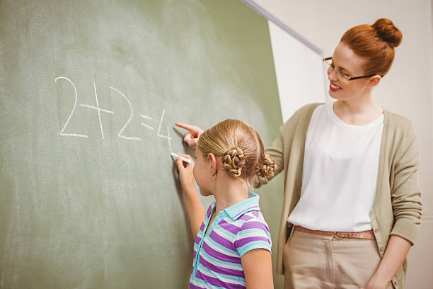 istock Teacher assisting girl to write on blackboard in classroom 474965390