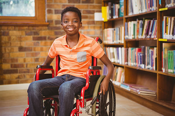 Imagem de uma criança centada em uma cadeira de rodas, é um menino de pele preta que está sorrindo ao seu fundo uma estante cheia de livros e uma parede de tijolos