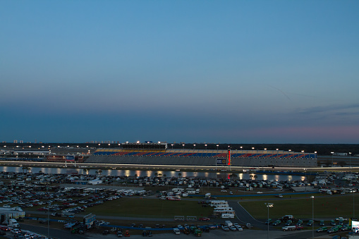 Daytona International Speedway at dusk before a NASCAR race in February of 2015. An orange and purple background sets the scene at the track. Lake Lloyd makes it's presence with the reflections of the lights reflected across it's surface.