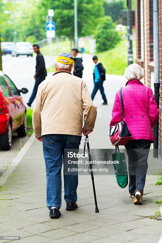 Caucasian senior couple walking along sidewalk Ratingen, Germany - May 16, 2015: Rearshot of a caucasian senior couple walking along sidewalk in Ratingen. Man is using a cane. He is wearing a cap. Woman is carrying to bags. In background three immigrant male people are walking. 2015 Stock Photo