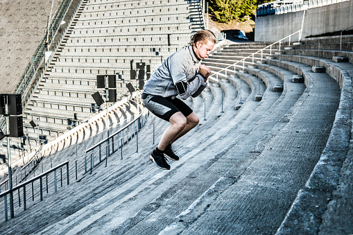 One man exercising inside an old stadium steps in early morning. He is doing squat exercise wearing gray professional sport clothing