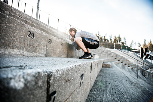 One man exercising inside an old stadium steps in early morning. He is doing squat exercise wearing gray professional sport clothing