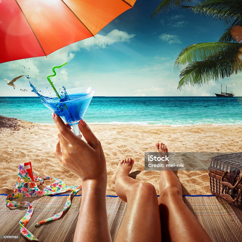 Vacation. Summer beach. Concept for rest. Close view Vacation. Summer tourism. Young woman is sitting on the sand with the blue cocktail in her hand Beach Stock Photo