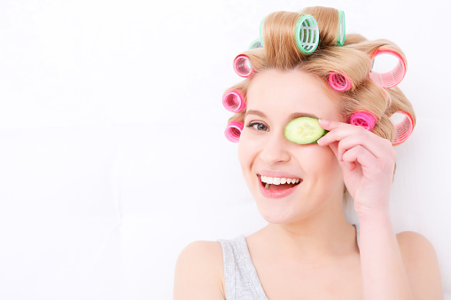 Beauty procedures. Portrait of a young beautiful blond girl sitting in the light background smiling and holding slice of cucumber near her eye while wearing pajama and colorful hair curlers 
