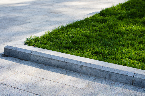 Green lawn in the flower bed in the Park surrounded by tile. Trimmed green grass illuminated by the rays of the sun. Flowerbed photographed as a triangle.