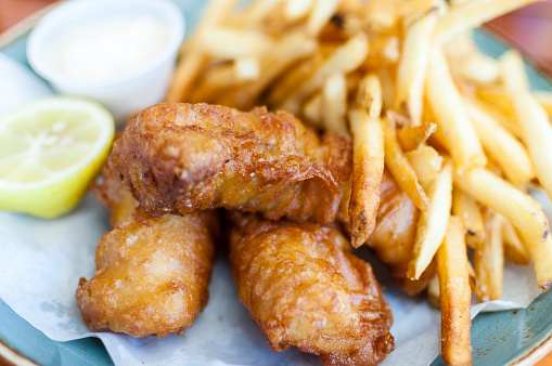 A close-up of delicious battered fish and chips/fries. Half a lemon and tartar sauce are also placed on the paper-covered plate, along side the fish and chips. A great image that can be used to capture: