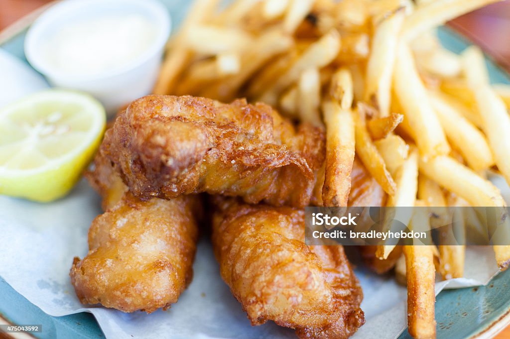 Fish and chips/fries on a plate A close-up of delicious battered fish and chips/fries. Half a lemon and tartar sauce are also placed on the paper-covered plate, along side the fish and chips. A great image that can be used to capture: Fish and Chips Stock Photo