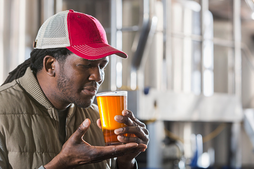 An African American man in a microbrewery, tasting a glass of craft beer with his eyes closed to savor the aroma, standing in front of storage tanks.  He is wearing a cap, long sleeves and a vest to stay warm in the chilled storage room.  He could be a worker or the owner of this small business.